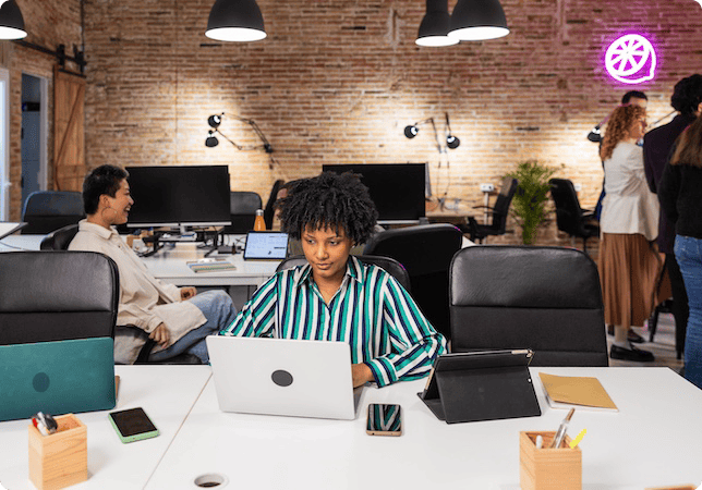 A woman sat at her desk in a busy workspace, working hard on her laptop.